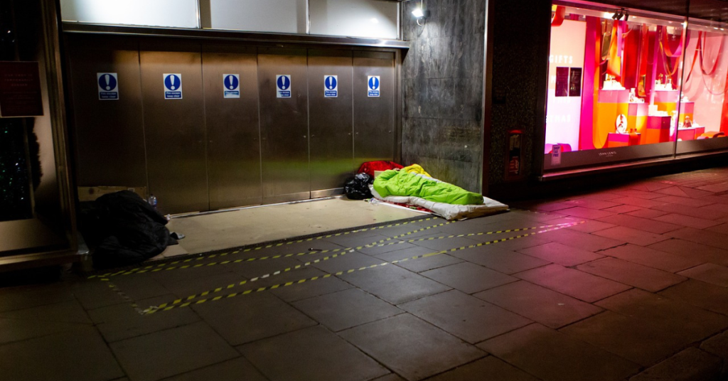 People sleeping rough in a doorway on a high street, Alongside, a brightly lit and decorated shop window displays expensive-looking trinkets