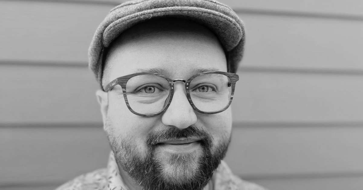 A close up black and white photo of Alex - a white man with a dark beard wearing glasses, a flat cap, and a shirt with leaves on it.