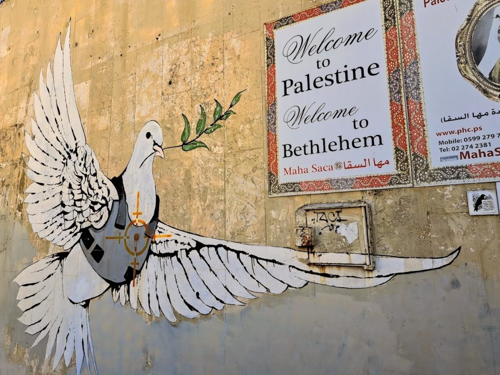 A dove wearing a bullet proof vest painted on a wall next to a sign saying Welcome to Palestine Welcome to Bethlehem