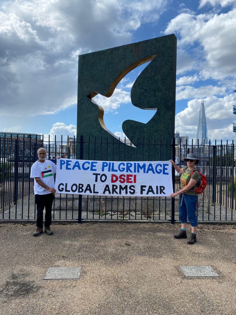 Two people holding up a banner reading 'Peace pilgrimage to DSEI global arms fair' standing in front of a memorial to victims of war with a backdrop of London