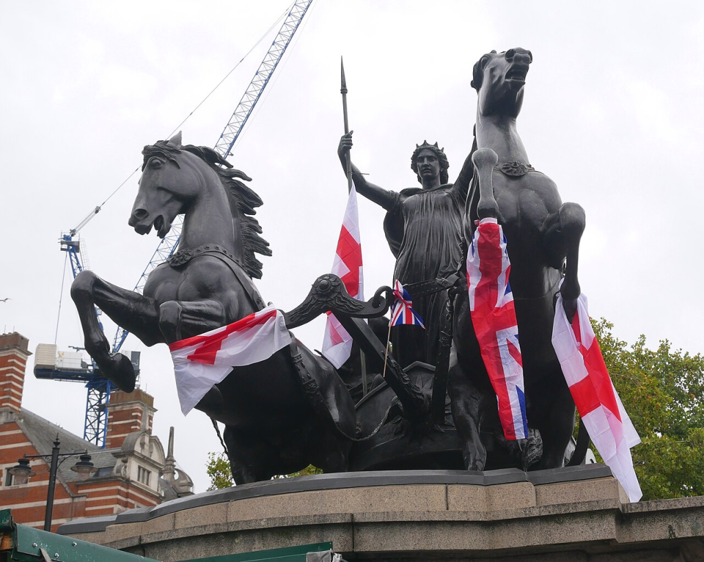 A statue of a chariot pulled by two rearing horses is draped with several English and UK flags, with construction cranes visible in the background.