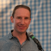 Simeon Mitchell in front of a microphone. In the background is a gingham checked backcloth inside a large marquee.